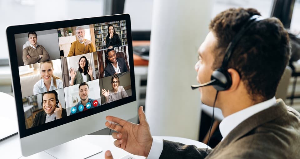 A man wearing a headset sits at a desk and speaks during a video conference on a computer screen, showing nine diverse people smiling and waving in individual windows. EFESO Management Consultants