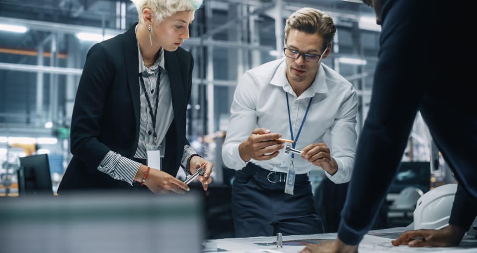 Two professionals in business attire collaborate at a table covered with documents and blueprints in a modern office setting. Both appear focused, discussing details and pointing at papers. EFESO Management Consultants