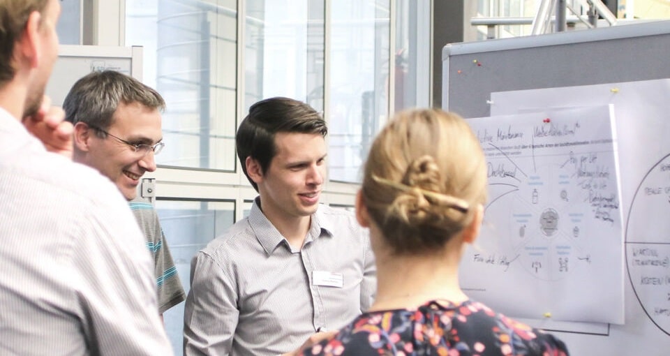 Four people stand in a group, discussing a diagram on a whiteboard in a bright, modern office setting. Two people face the board, while two others listen and engage in conversation. EFESO Management Consultants