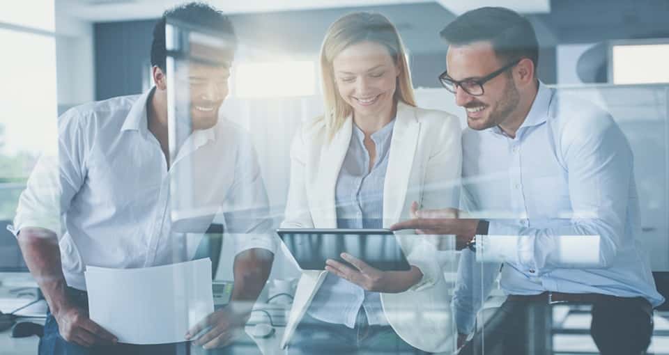 Three business professionals, two men and one woman, are smiling and looking at a tablet together in a bright modern office setting. One man holds papers, and the group appears to be collaborating. EFESO Management Consultants