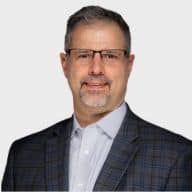A middle-aged man with short gray hair, glasses, and a goatee, wearing a checked suit jacket and light-colored collared shirt, posing against a plain white background. EFESO Management Consultants