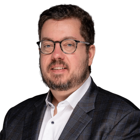 A man with short dark hair, glasses, and a beard, wearing a white shirt and a dark plaid blazer, smiling at the camera against a plain light background. EFESO Management Consultants