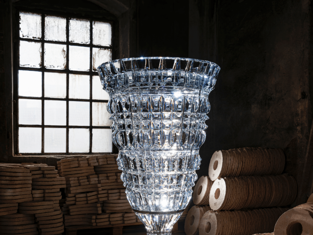 A large, intricately cut glass vase stands in a dimly lit workshop, with stacks of wooden disks and an old, multi-pane window in the background. EFESO Management Consultants