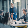 Four people are having a business meeting in a modern office. Three are seated at a table with laptops and documents, while one person, standing and smiling, appears to be leading the discussion. EFESO Management Consultants